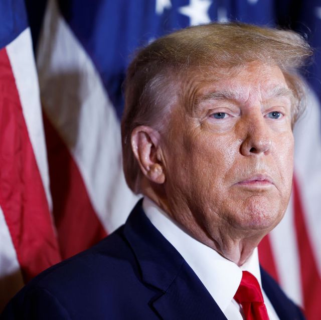 donald trump stands in front of several american flags that are out of focus in the background, he looks off camera to the right with a neutral expression on his face, he is wearing a navy blue suit jacket, white collared shirt and red tie