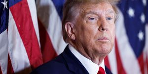 donald trump stands in front of several american flags that are out of focus in the background, he looks off camera to the right with a neutral expression on his face, he is wearing a navy blue suit jacket, white collared shirt and red tie