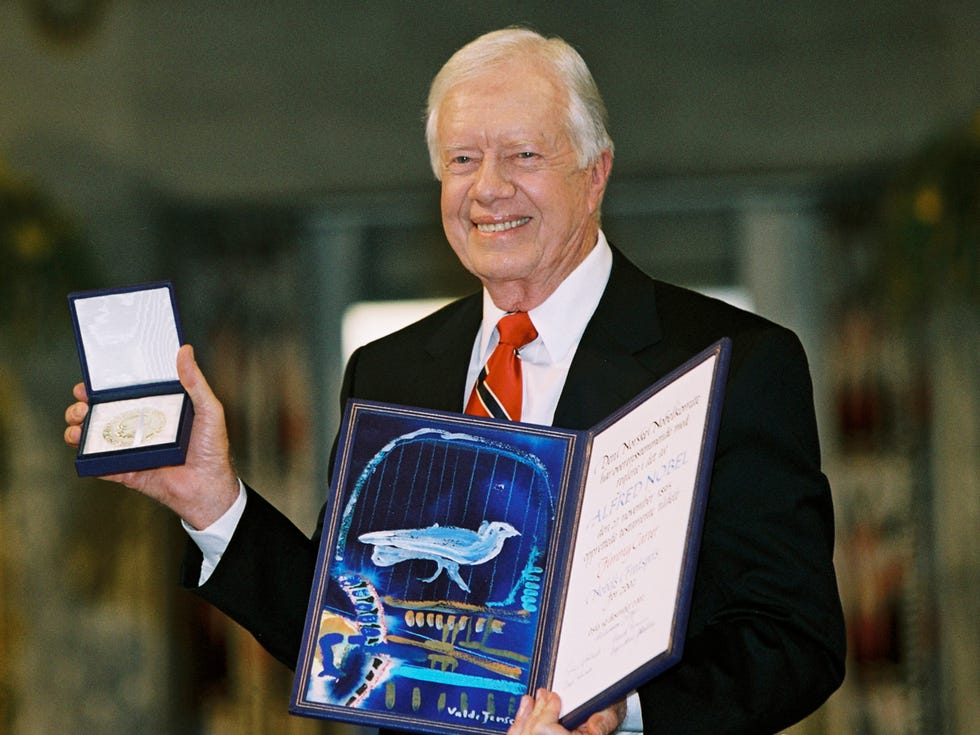 jimmy carter accepts the nobel peace prize in 2002, he holds a small box with the medal inside in his right hand, in his left hand, he holds a certificate for the prize that includes a blue, white and gold drawing, carter is smiling and wearing a black suit jacket, white collard shirt and red tie with a black and white stripe