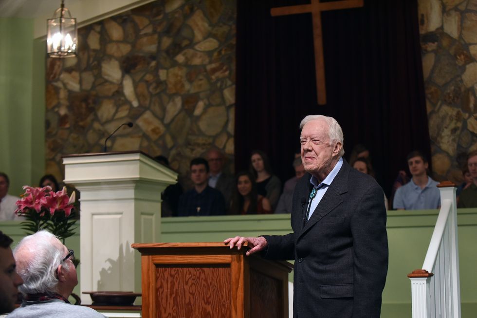 jimmy carter stands next to a wooden podium and smiles as he looks forward, he wears a dark suit jacket and blue collared shirt, people are sitting in front of and behind him
