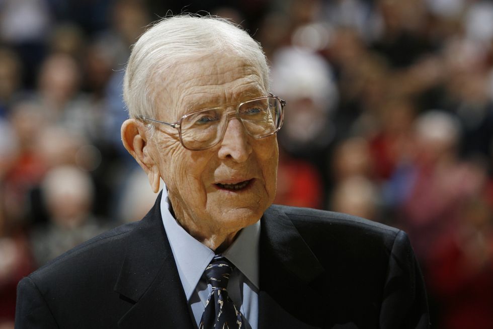 john wooden wearing a black suit and blue shirt, and glasses, smiling and looking off camera at a basketball game