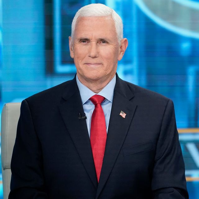 mike pence smiles at the camera while sitting in a white high backed chair, he wears a dark suit jacket, blue collared shirt and red tie with an american flag pin on his lapel