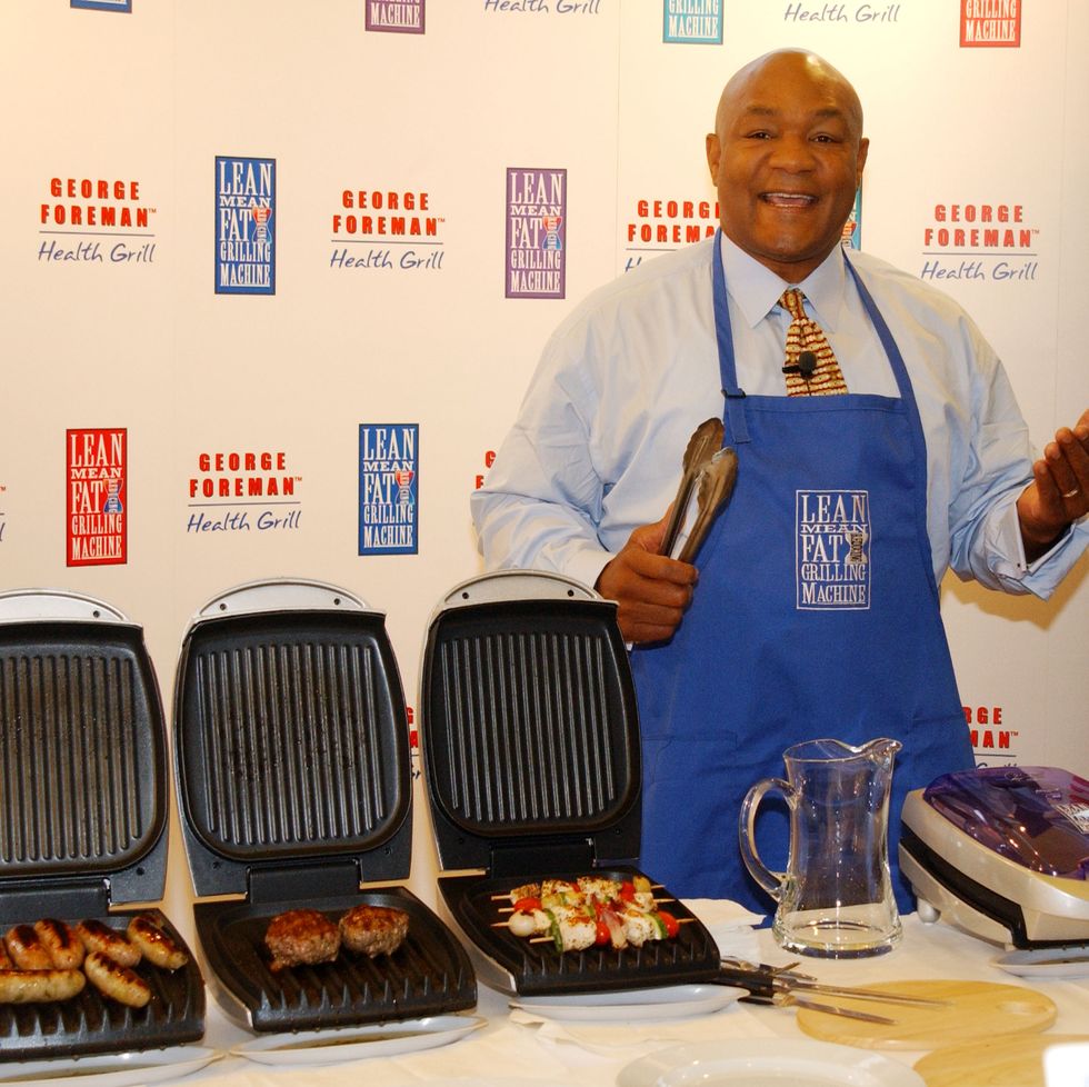 george foreman smiling as he stands behind a table of grills
