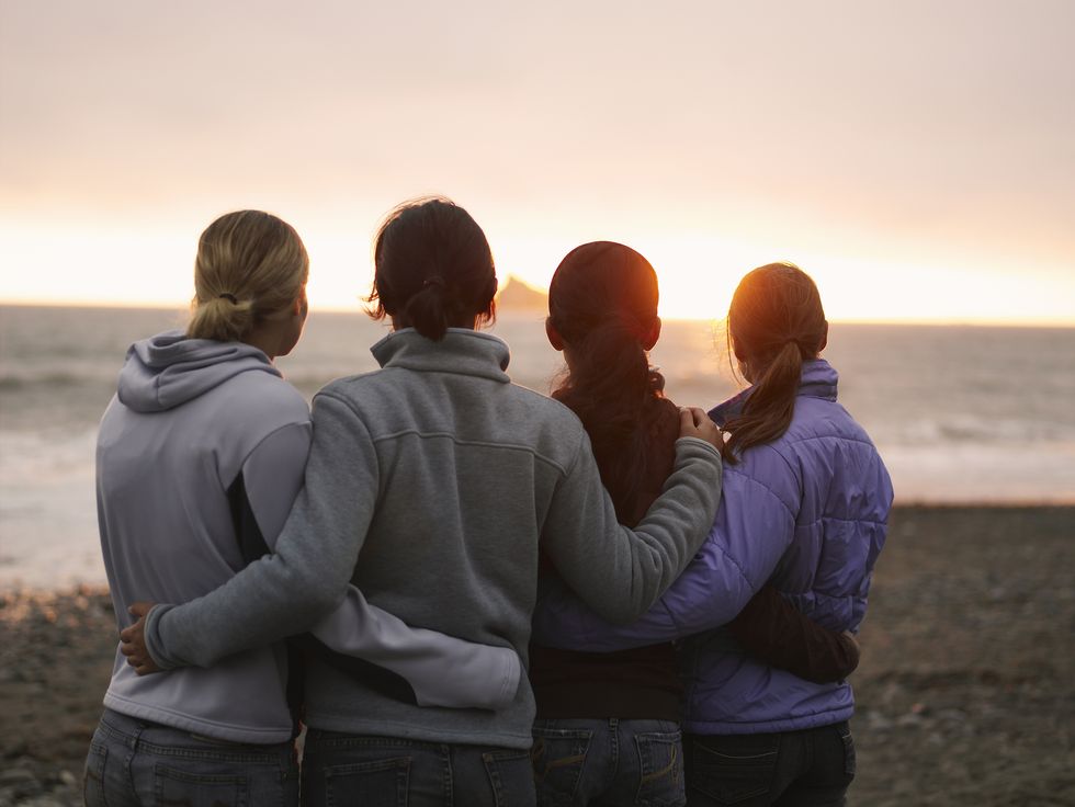 four young women, arms around each other, looking out to sea, rear view
