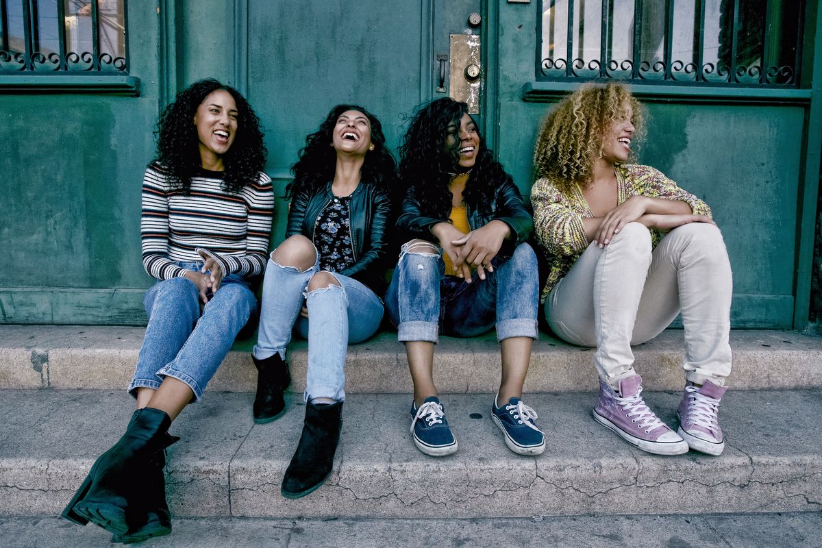 Four young women with curly hair sitting side by side on steps outside a building.