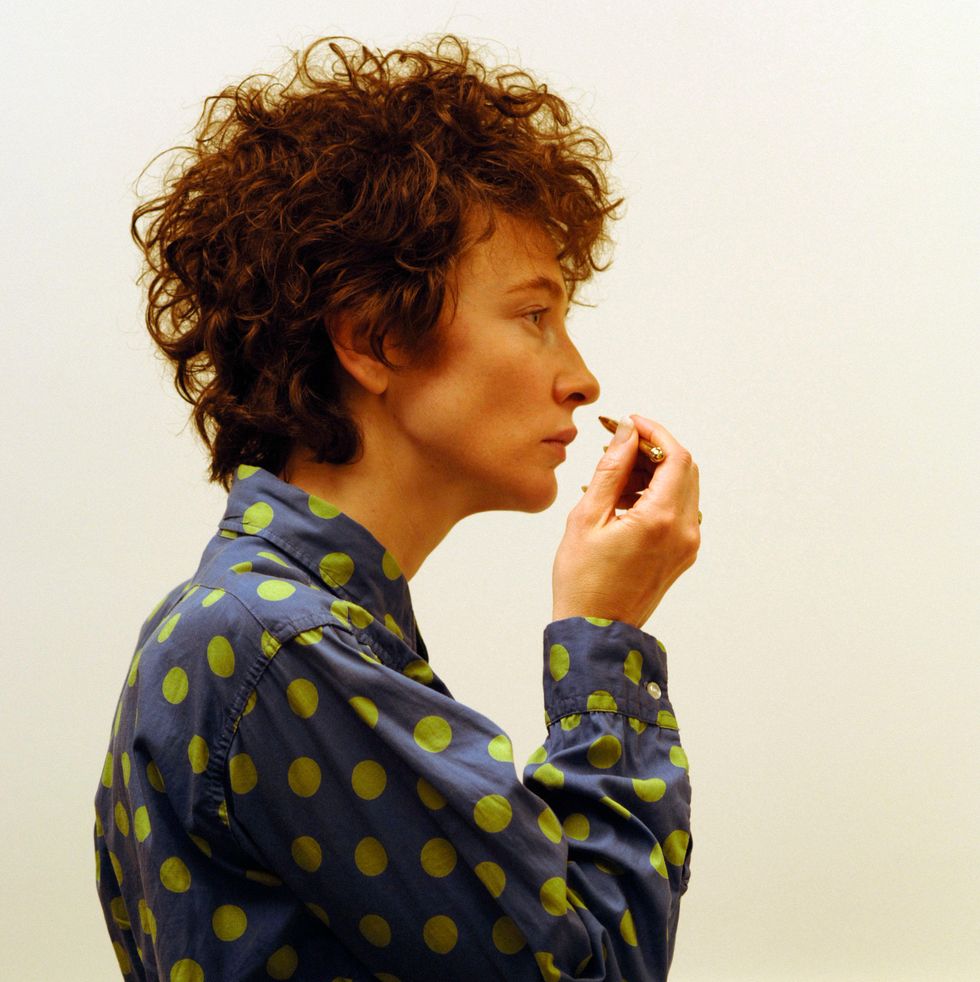 cate blanchett in character as bob dylan wearing a blue collared shirt with green polka dots, she stands in profile and raises a short pencil to her face
