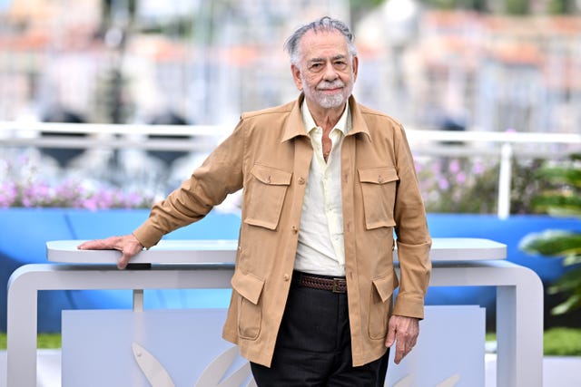 francis ford coppola smiles at the camera as he stands outside wearing a tan jacket and a light colored collared shirt that is unbuttoned at the neck