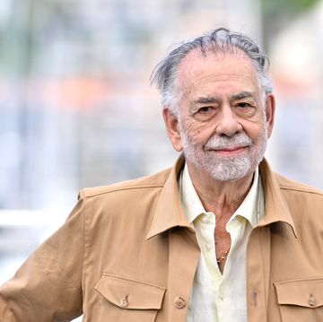 francis ford coppola smiles at the camera as he stands outside wearing a tan jacket and a light colored collared shirt that is unbuttoned at the neck