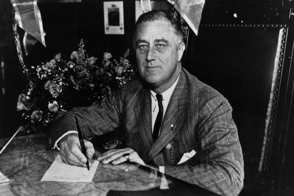 franklin roosevelt wearing a suit and tie, sitting at a table, signing a piece of paper with a pen