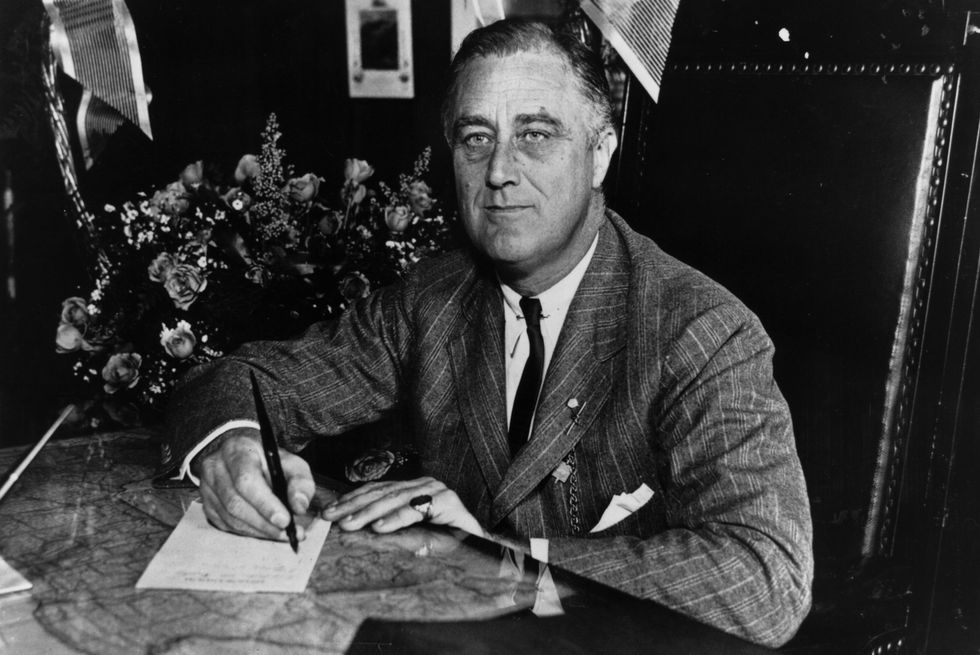 franklin roosevelt wearing a suit and tie, sitting at a table, signing a piece of paper with a pen