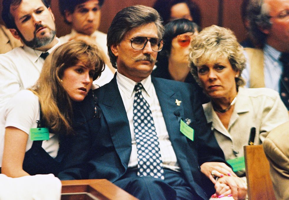 kim goldman, fred goldman, and patti goldman sit on a bench and look ahead, both women lean into fred as the family holds hands and embraces, many people sit behind