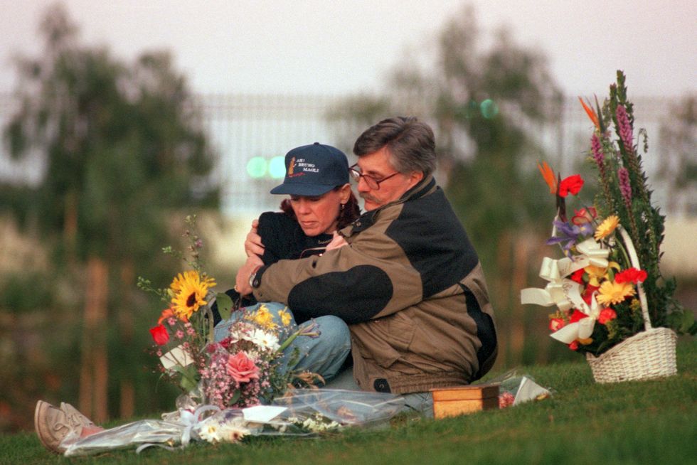 patti and fred goldman sit on grass and look down at the ground, in front of them are several flower bouquets, fred hugs patti
