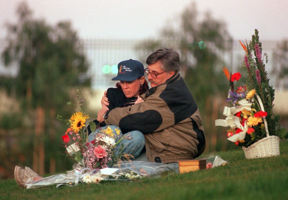 patti and fred goldman sit on grass and look down at the ground, in front of them are several flower bouquets, fred hugs patti