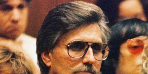 fred goldman looks to the left, he wears aviator glasses, a blue suit jacket and a patterned shirt and tie