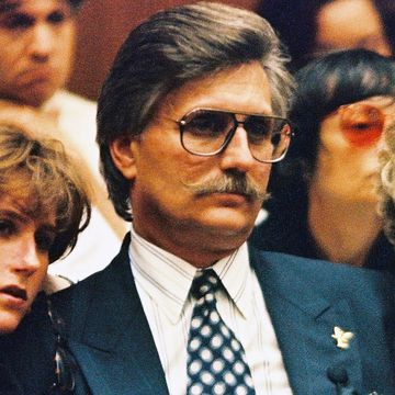 fred goldman looks to the left, he wears aviator glasses, a blue suit jacket and a patterned shirt and tie