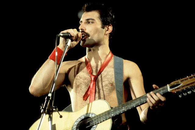 freddie mercury stands on a stage with a dark backdrop, speaking into a microphone on a stand, wearing no shirt, holding a guitar