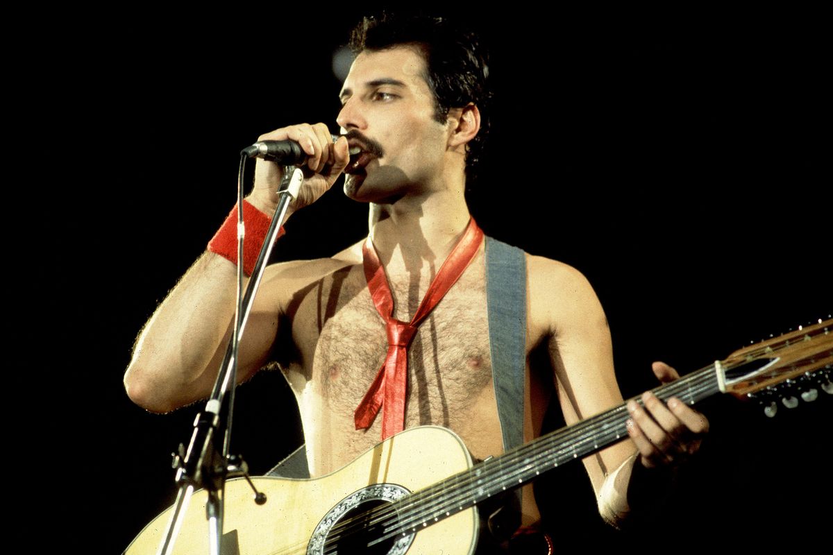 freddie mercury stands on a stage with a dark backdrop, speaking into a microphone on a stand, wearing no shirt, holding a guitar