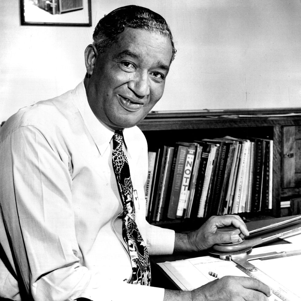 frederick mckinley jones smiles at the camera while sitting at a table with a pencil in hand, paper and equipment rests on the table in front of him, behind is a bookshelf, he wears a collared shirt and patterned tie