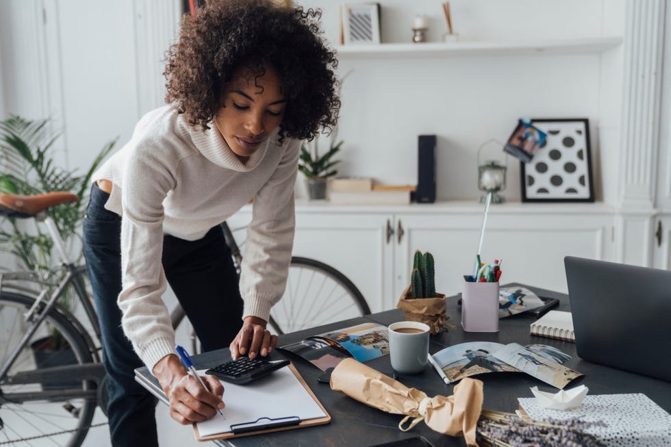 jobs for stay at home moms - Woman of color standing at her desk, using calculator, taking notes