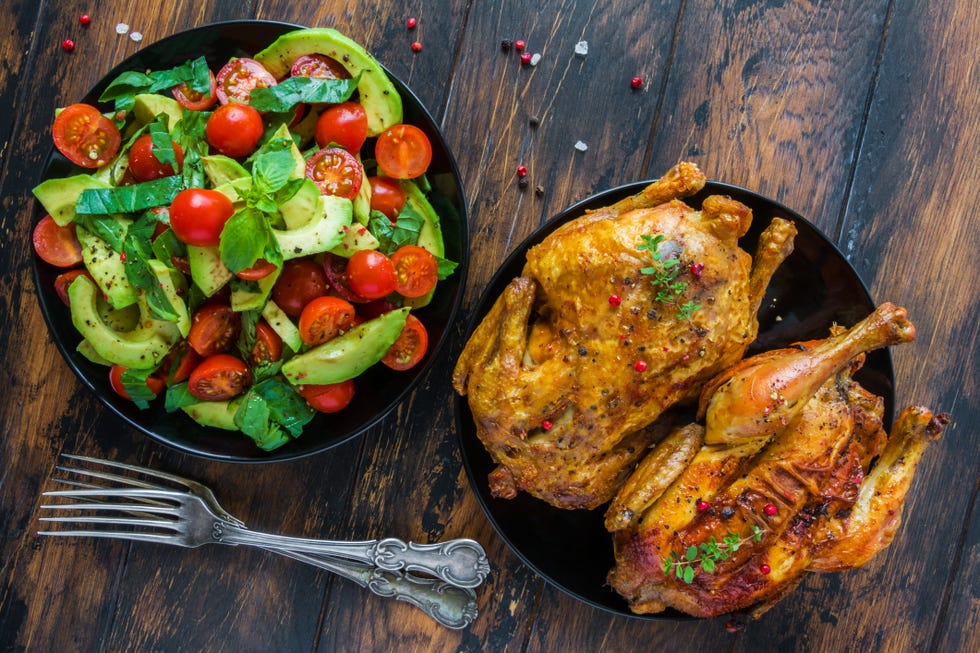 French crispy chicken baked with garlic and thyme and salad with avocado, basil leaves and cherry tomatoes. Black plates on the wooden rustic table, top view.