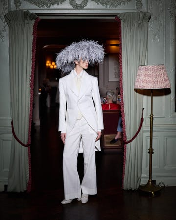 A model wearing a white tailored suit and an elaborate feathered headpiece stands in a doorway