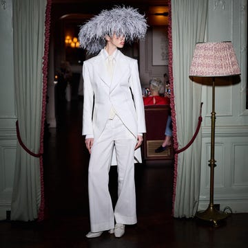 A model wearing a white tailored suit and an elaborate feathered headpiece stands in a doorway A model wearing a white tailored suit and an elaborate feathered headpiece stands in a doorway