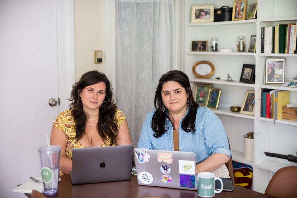 two individuals are seated at a wooden table, focused on their laptops one person wears a yellow floral top and the other, a light blue shirt