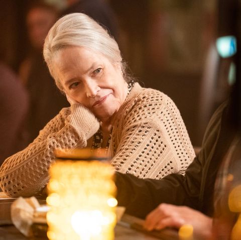 a woman leans her head against her palm as she looks to the right, she wears a tan sweater and sits at a bar