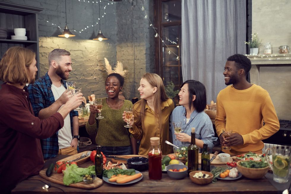 multi ethnic group of smiling young people enjoying dinner together standing at table in modern interior and holding wine glasses