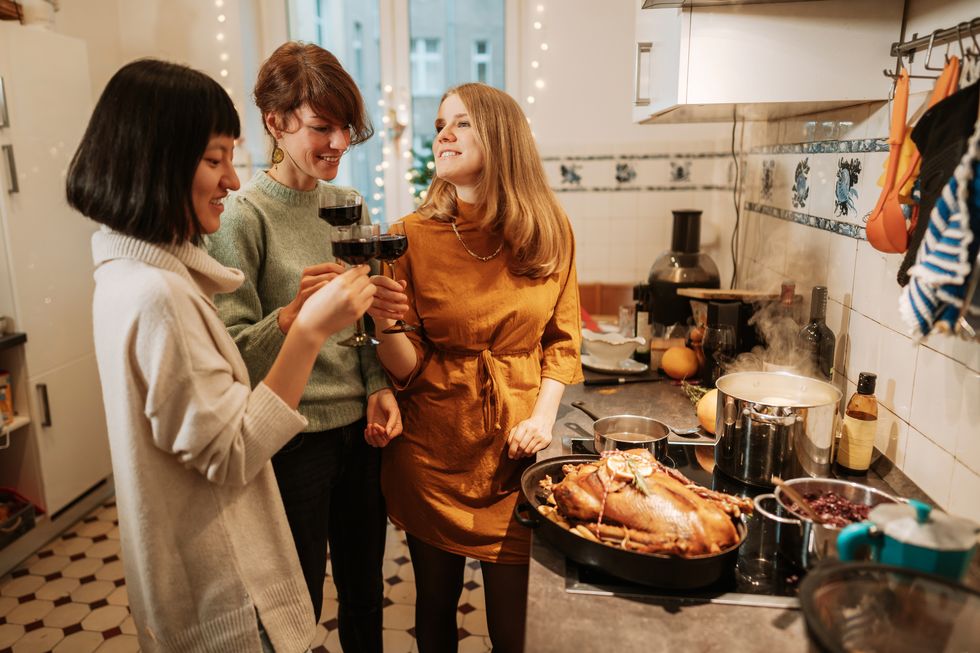 three women drinking red wine while preparing turkey meal at home with a turkey and three pot on the stove cooking