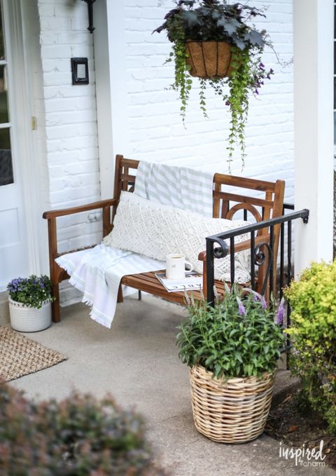 front porch with plants in pots and a wood bench with long white throw pillow and white throw with a magazine and cup of coffee sitting on it