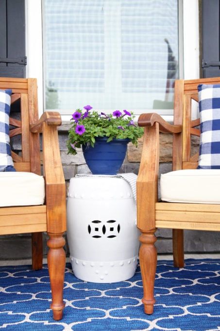 close up of two wooden chairs with white seat cushions and blue and white plaid pillows on top of a blue and white rug with a white ceramic garden stool and blue pot with flowers between the chairs