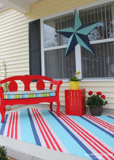 front porch with red bench with colorful patterned cushion, red crate as a side table with a plant in a yellow pot on it and a diagonally striped rug in red, blue and white
