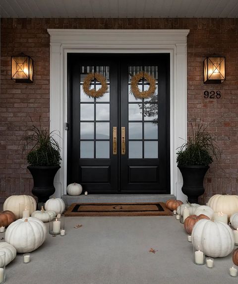 doorway of house with white yellow and orange pumpkins and lit candles lining walkway and steps there is a welcome mat with the monogram g on it, two large planters on either side of the door and two wheat wreaths on the black double doors