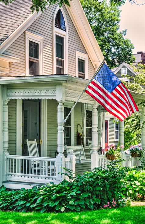 a backlit american flag waves over a front porch of a cape cod home there are white wood rockers and a white wood swing on the porch and flower boxes in front of