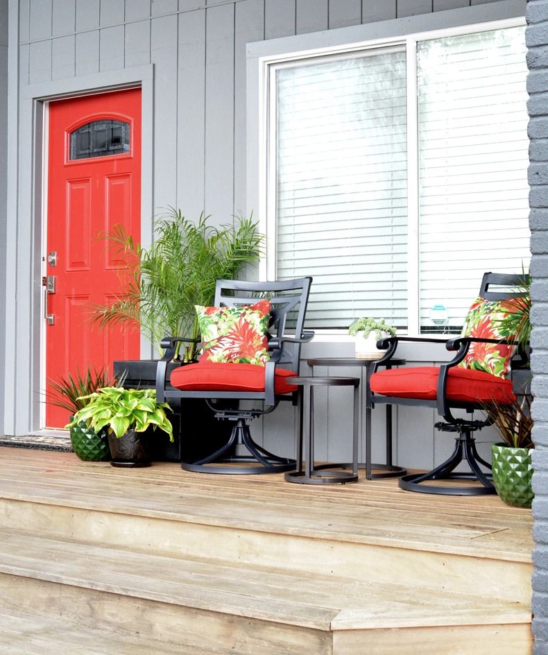 wood floor front porch with two black metal chairs with red cushions and tropical plant printed pillows and two black metal nesting tables between them there are tropical plants on the porch and a red front door