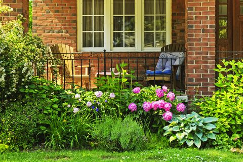 front of home with two adirondack chairs on front porch and flower garden in front with flowering peony bush