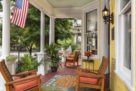 front porch of yellow house with american flag in summer with two seating areas one with a patterned rug and two wood chairs with orange cushions facing each other, the other with two chairs looking out from the porch side by side