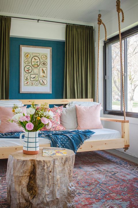 bed swing with pink and white pillows and a blue throw hanging on an enclosed porch over a distressed oriental style rug with a table made from a tree stump with a pitcher of flowers on it