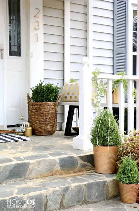 stone front porch with two steps with plants in gold pots, a gold polka dot white pillow on a white and black bench, a plant in a wicker basket and a striped black and white doormat