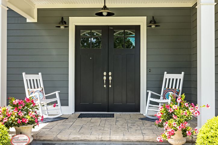 a front porch with two white rocking chairs with patterned pillows, stamped concrete floors, pots with pink flowers and double glass doors