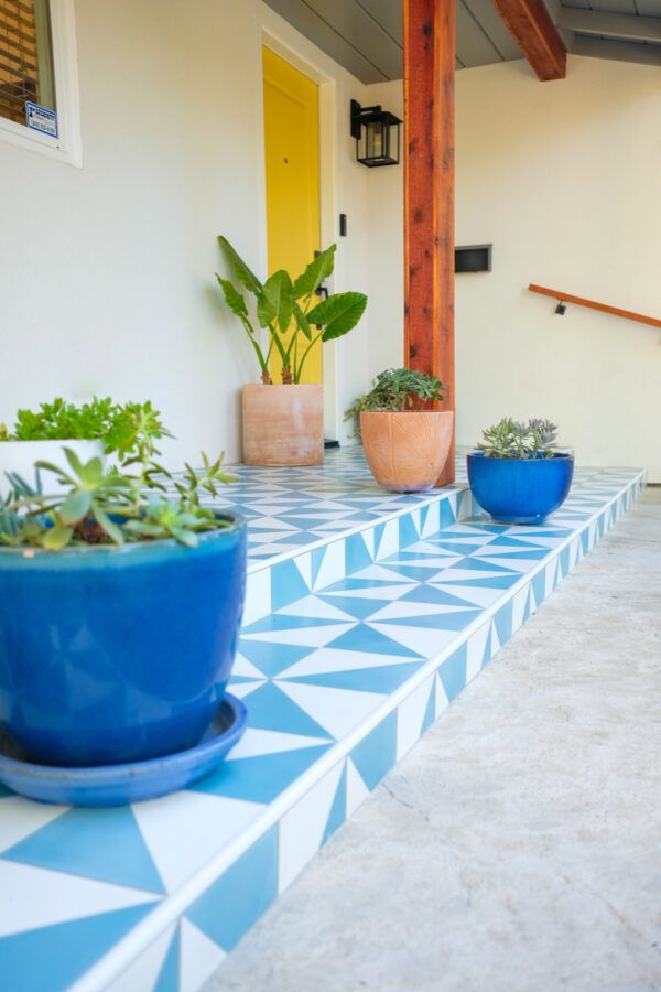 front porch with white and light blue geometric patterned tile floor with pots of plants sitting on it and a yellow front door