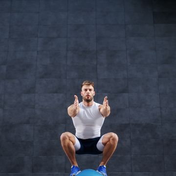front view of handsome caucasian muscular bearded man doing squat exercise on bosu ball in background is gray wall