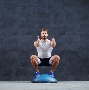 front view of handsome caucasian muscular bearded man doing squat exercise on bosu ball in background is gray wall
