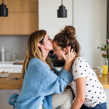 front view portrait of mature mother with adult daughter sitting at home, kissing forehead