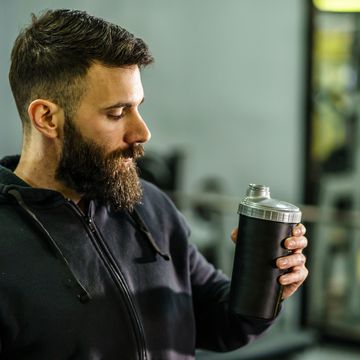 front view portrait of young caucasian man athlete in black hoodie male standing in the gym holding protein supplement shaker supplementation in training waist up black hair and beard copy space drink