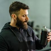 front view portrait of young caucasian man athlete in black hoodie male standing in the gym holding protein supplement shaker supplementation in training waist up black hair and beard copy space drink