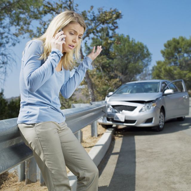 frustrated woman using cell phone next to car wrecked on guardrail