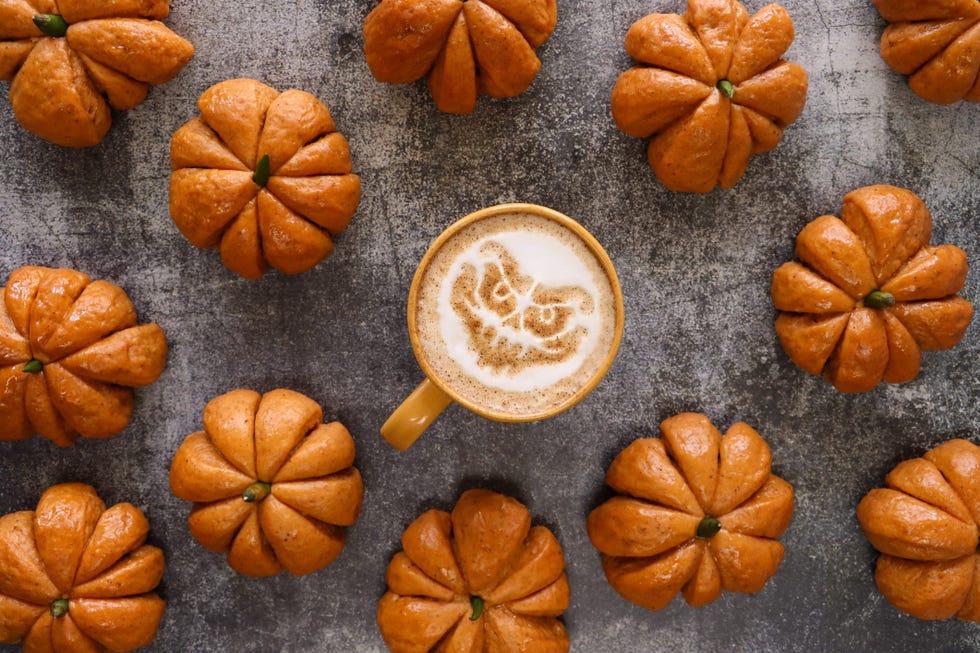 full frame image of orange coffee mug containing latte with halloween milk froth art design of scary ghoul face surrounded by pumpkin designed rolls with green chilli pepper stalks, homemade halloween bread buns glazed with apricot jam, elevated view