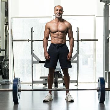 full length of shirtless man with barbell standing in gym